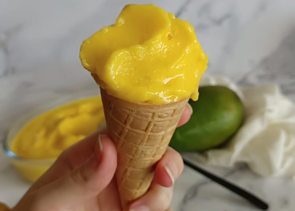 Foto de la receta de helado de mango saludable de Nitka fitness. La imagen muestra el resultado de la receta, mostrando la cremosidad del helado encima de una galleta en forma de cono, con la tarrina de helado de mango de fondo.