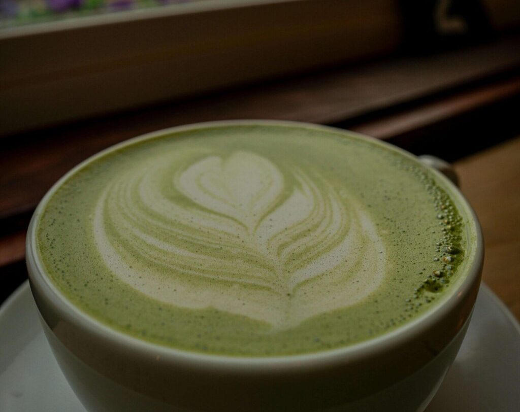 Imagen de una escena tranquila y serena: una taza blanca de té matcha recién preparado reposa delicadamente ante una ventana iluminada, permitiendo la luz realce su tonalidad verde vibrante. 